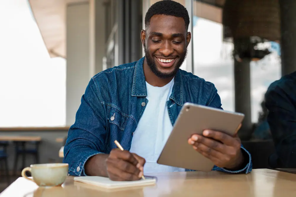Smiling young man sitting at a table, using a tablet and writing in a notebook.